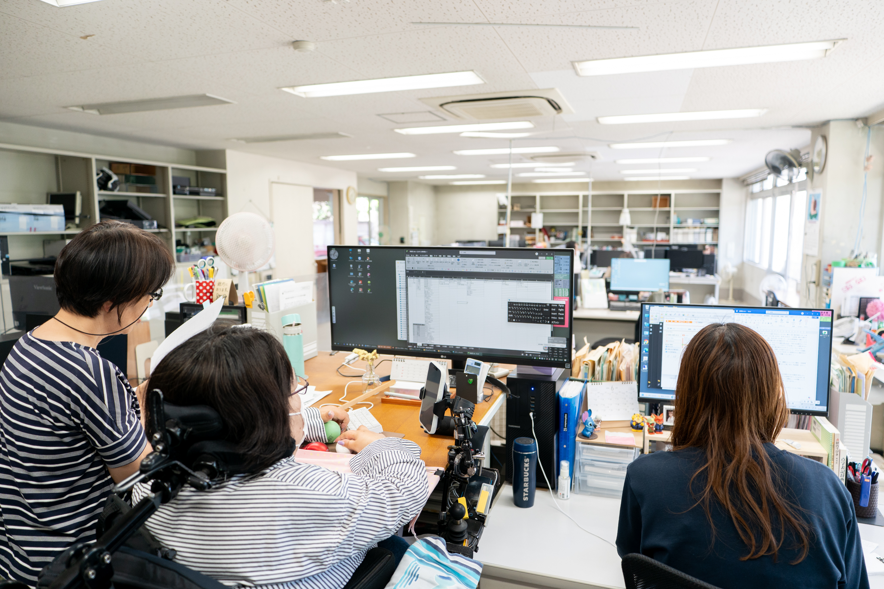 【写真】高めのデスクで作業をするひとと、そのそばで書類を眺めるひと