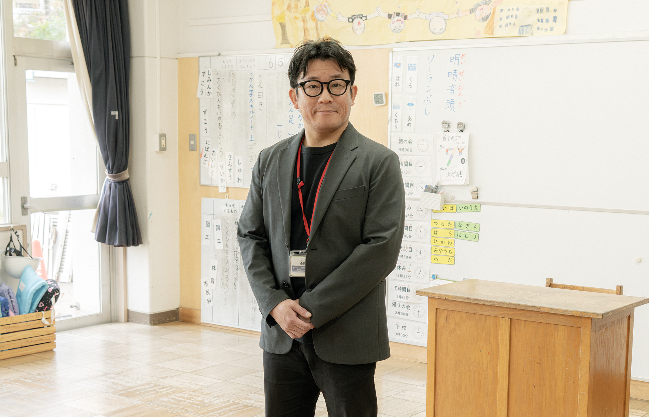 【写真】小学校の教室のような場所。黒いインナーにグレーのジャケットを着ているおのさんが立っている。黒ふちメガネをしている。