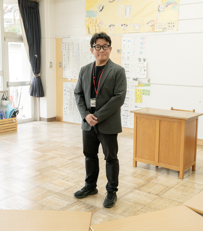 【写真】小学校の教室のような場所。黒いインナーにグレーのジャケットを着ているおのさんが立っている。黒ふちメガネをしている。