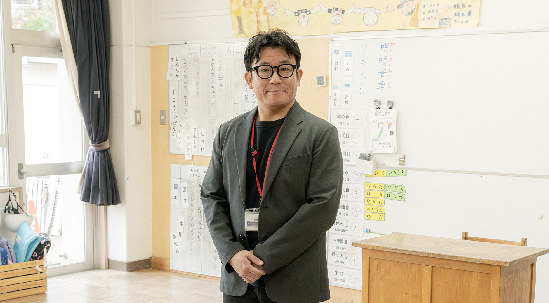 【写真】小学校の教室のような場所。黒いインナーにグレーのジャケットを着ているおのさんが立っている。黒ふちメガネをしている。
