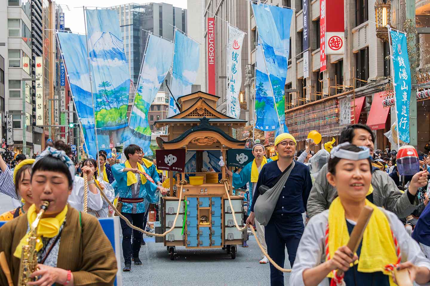 作品《銭湯山車》を引きながら、音楽を奏でたり、幟を立てたりする人々が街を巡行している様子。