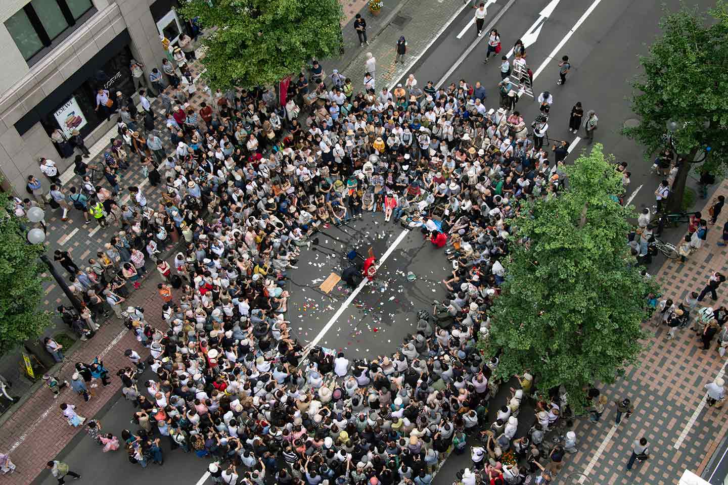 ギリヤーク尼ヶ崎さんの路上大道芸を上空から撮影した写真。大勢のギャラリーが路上に座り込み、その中央には円形の空白があり、ギリヤーク尼ヶ崎さんがパフォーマンスを行っている。