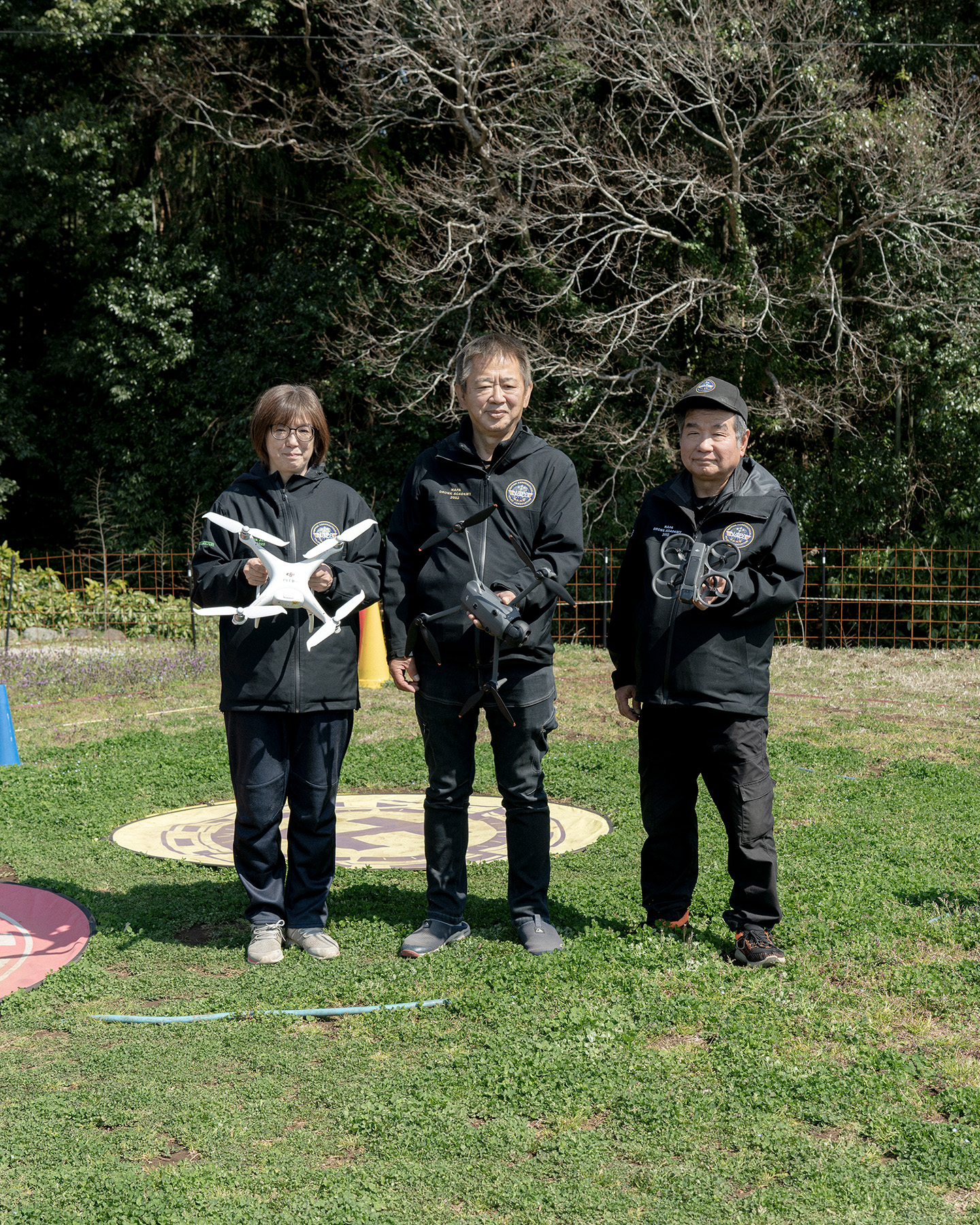 【写真】屋外に上下黒色のユニフォームを着た三人が立っている。一人ずつ手には異なる型のドローンを持ち、正面を向いている。足元は芝生で、ドローンを止めるための丸いマークがついている。