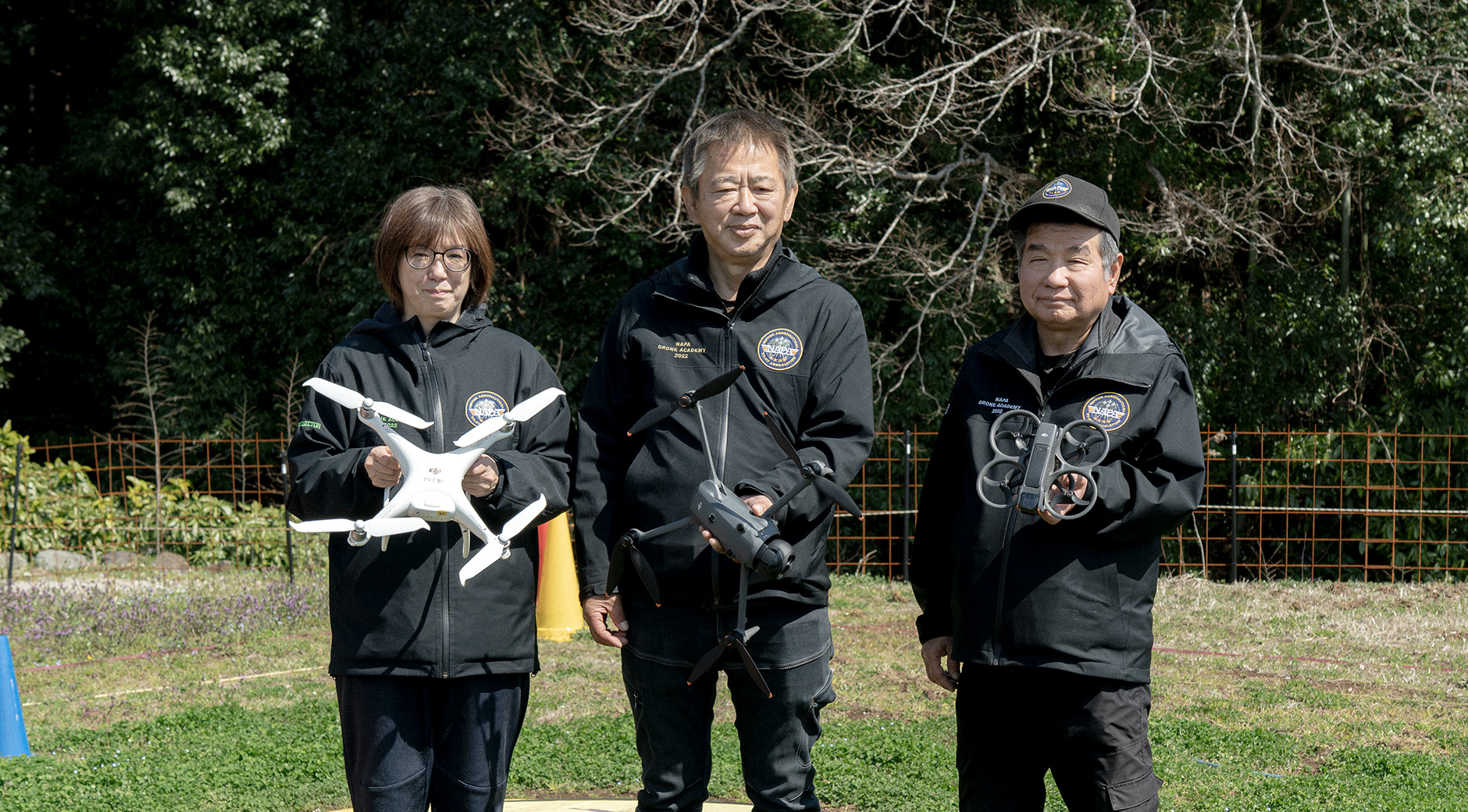 【写真】屋外に上下黒色のユニフォームを着た三人が立っている。一人ずつ手には異なる型のドローンを持ち、正面を向いている。足元は芝生で、ドローンを止めるための丸いマークがついている。