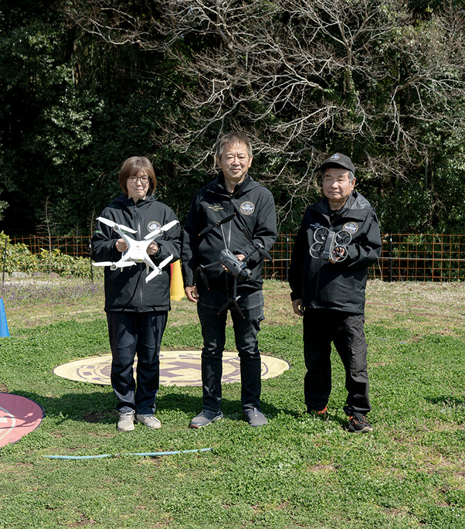 【写真】屋外に上下黒色のユニフォームを着た三人が立っている。一人ずつ手には異なる型のドローンを持ち、正面を向いている。足元は芝生で、ドローンを止めるための丸いマークがついている。