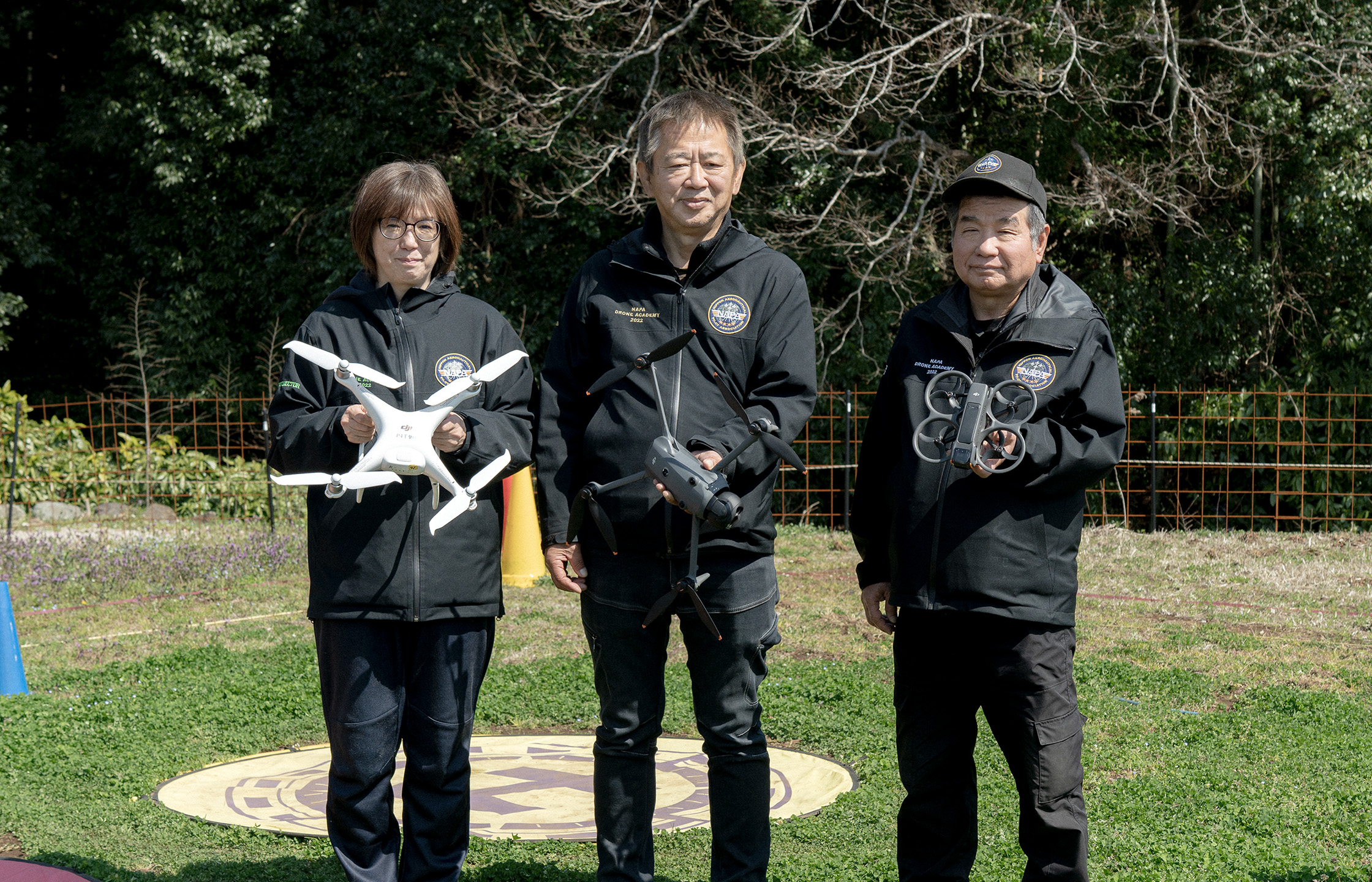 【写真】屋外に上下黒色のユニフォームを着た三人が立っている。一人ずつ手には異なる型のドローンを持ち、正面を向いている。足元は芝生で、ドローンを止めるための丸いマークがついている。